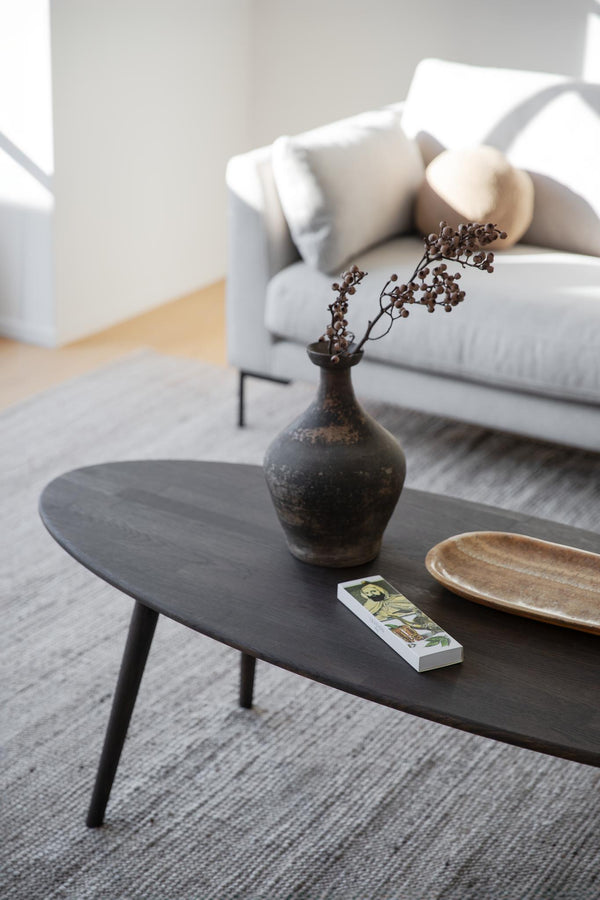Mörkt ovalt soffbord i trä med keramikvas, träskål och dekorativa grenar i minimalistiskt vardagsrum