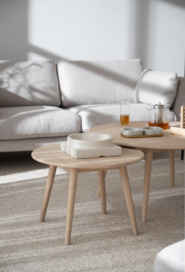 Ljusa soffbord i trä med rund bordsskiva och tre ben, dekorerade med keramikskålar och tekanna framför beige soffa