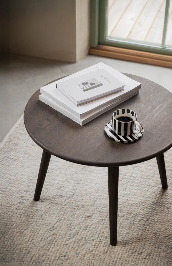 Runt soffbord i mörk ek med böcker och randig kaffekopp på ljus matta vid fönster