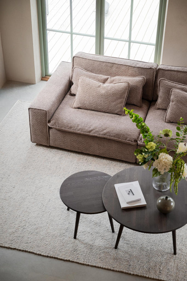 Beige boucle-soffa med kuddar vid fönster, mörkgrå soffbord och beige matta i minimalistiskt vardagsrum