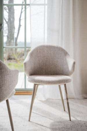 Grå stoppad stol med armstöd och träben framför fönster med vit gardin i ljust rum
