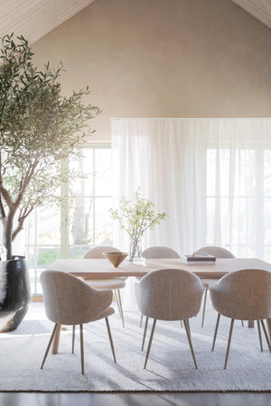 Ljus matplats med oval matbord i trä och beige stolar, stora fönster med vita gardiner och olivträd