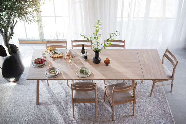 Matbord i ljust trä med iläggsskiva och sex stolar i modern skandinavisk design med vita gardiner i bakgrunden