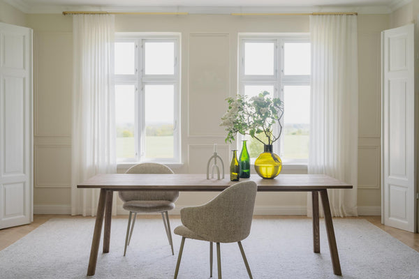 Matbord i trä med stolar vid fönster i ljust matrum med vita gardiner och dekorativa vaser
