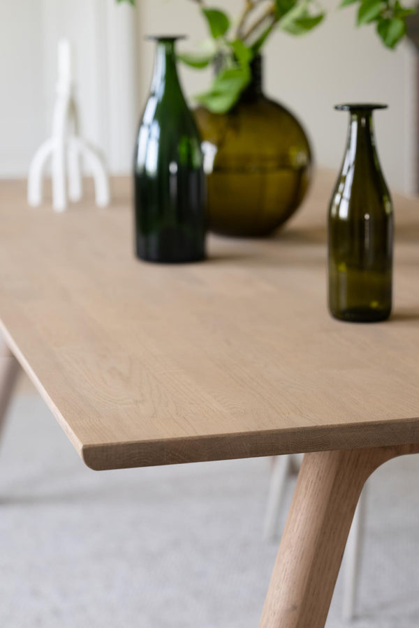 Ljust träbord med dekorativa glasflaskor i grönt, vit vas och gröna växtdetaljer