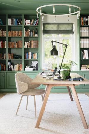 Hemmakontor med gröna bokhyllor, skrivbord i ljust trä, beige stol och modern lampring i taket