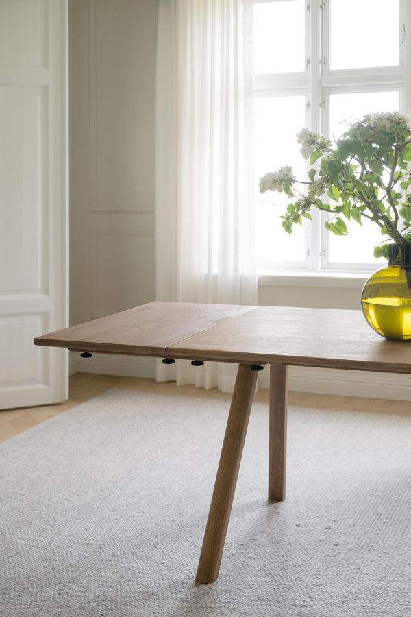 Utdragbart matbord i ljust trä med snedställda ben i skandinavisk design, placerat i ljust rum med fönster