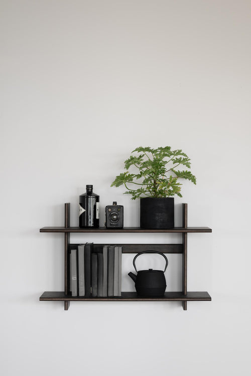 Minimalistisk väggmonterad hylla i mörkt trä med böcker, växt, kamera och tekanna mot vit vägg