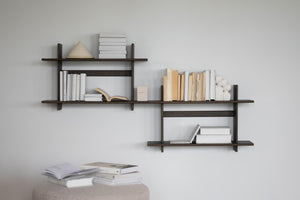 Väggmonterad bokhylla i mörkt trä med asymmetrisk design, fylld med böcker och dekorationsobjekt mot ljusgrå vägg