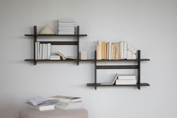 Väggmonterad bokhylla i mörkt trä med asymmetrisk design, fylld med böcker och dekorationsobjekt mot ljusgrå vägg