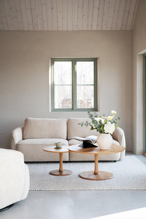 Ljust vardagsrum med beige soffa, två runda träsoffbord och vita blommor vid fönster med grå väggar