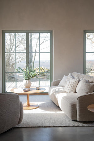 Ljust vardagsrum med beige soffa, rund soffbord i trä och utsikt genom altandörr mot natur