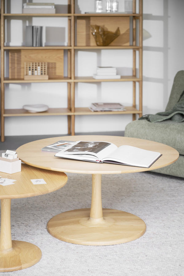 Runt soffbord i ljust trä med pedestal och öppen bok, bredvid grön fåtölj och bokhylla i skandinavisk stil