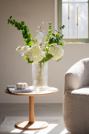 Vit vas med konstgjorda blommor och gröna blad på runt sidobord i modernt vardagsrum