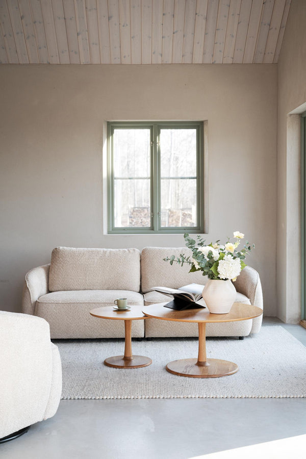 Ljust vardagsrum med beige soffa, runda träsoffbord och vit vas med blommor framför fönster