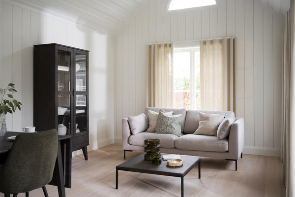 Ljust vardagsrum med beige soffa, svart soffbord, vitrinskåp och ljusa gardiner i skandinavisk stil