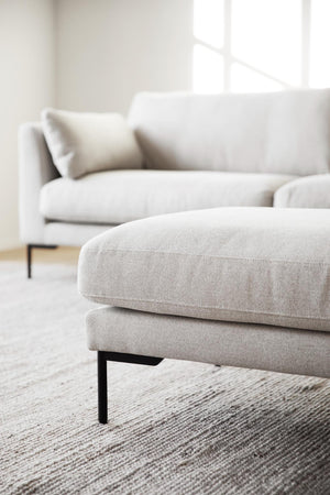 Beige ottoman med mjuk klädsel och svarta metallben framför matchande soffa i ljust vardagsrum