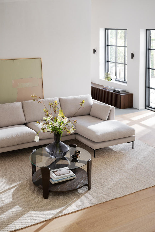 Modernt vardagsrum med beige hörnsoffa, runt soffbord i glas och mörkt trä, ljusa väggar och svarta fönster