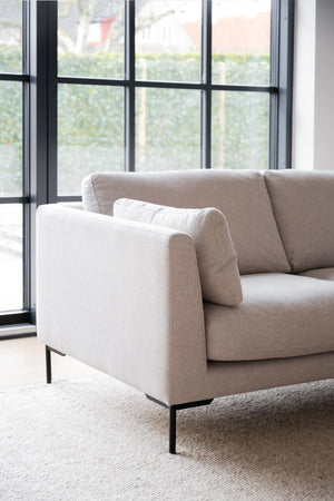 Beige soffa med svarta metallben framför stora industrifönster i ljust vardagsrum