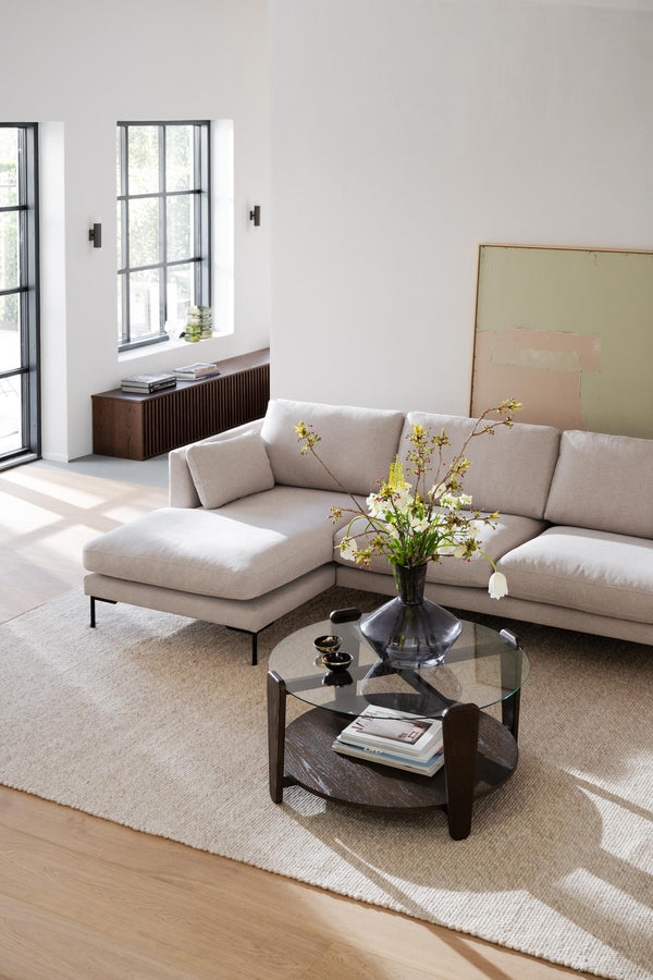 Ljust vardagsrum med beige hörnsoffa, runt soffbord i glas och mörkt trä, och minimalistisk inredning