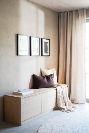 Ljus hallmöbel i trä med förvaringsluckor, dekorerad med kuddar och pläd vid fönster med gardiner