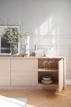 Ljust trä sideboard med öppna hyllor, dekorerat med vaser, böcker och glasservering mot vit vägg