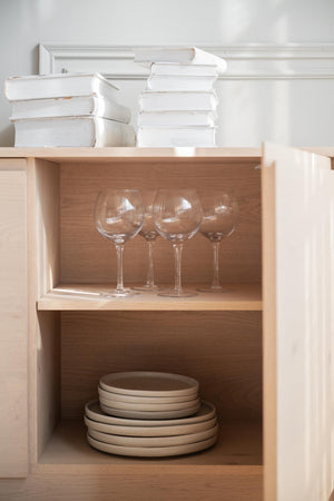 Öppen skåpsektion i ljust trä med glasvin och staplade tallrikar i beige keramik