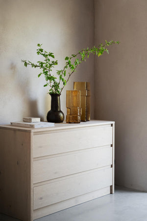 Ljus träbyrå med tre lådor i ljust trä, dekorerad med svart vas med gröna kvistar och gyllene glasvaser