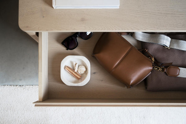 Öppen låda med brunt läderväska, solglasögon och småförvaring i vit skål på träbord