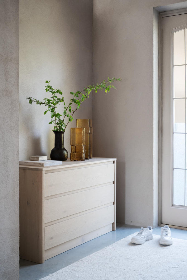 Ljus träbyrå med tre lådor i minimalistisk hall med grönväxter och guldfärgade vaser