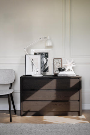 Mörk träbyrå med tre lådor, dekorerad med tavlor, lampa och dekorationer i skandinavisk stil