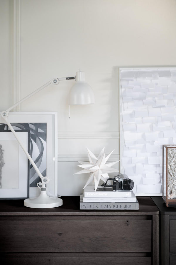 Vit skrivbordslampa med justerbar arm bredvid dekorativa föremål och inramade bilder på mörkt träbord