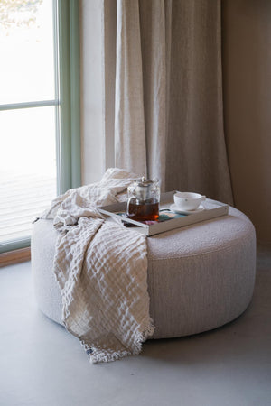 Beige pall i bouclé med vit filt, tekopp och tekanna vid fönster med linnegardin