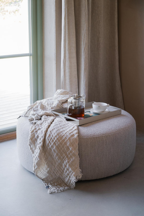 Beige pall i bouclé med vit filt, tekopp och tekanna vid fönster med linnegardin