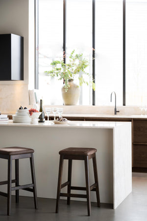 Modernt kök med vit köksö, mörka barstolar och stor fönstervägg med grönväxter