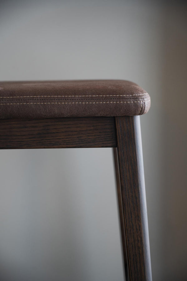 Närbild på stoppad sits i brunt konstläder med synliga sömmar på pall eller stol med mörkt trä