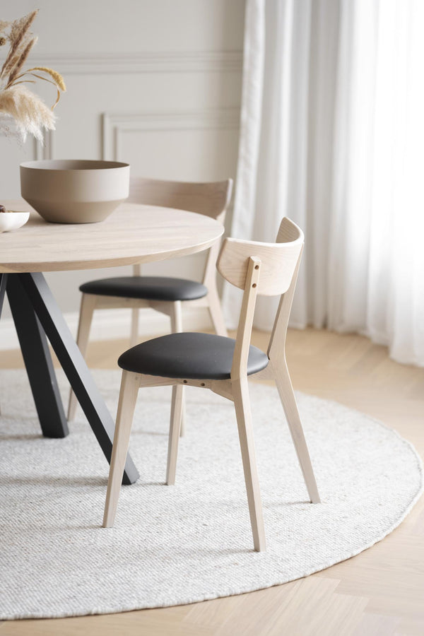 Rund matbord i ljust trä med svarta ben och två stolar med svarta dynor i minimalistisk matsal