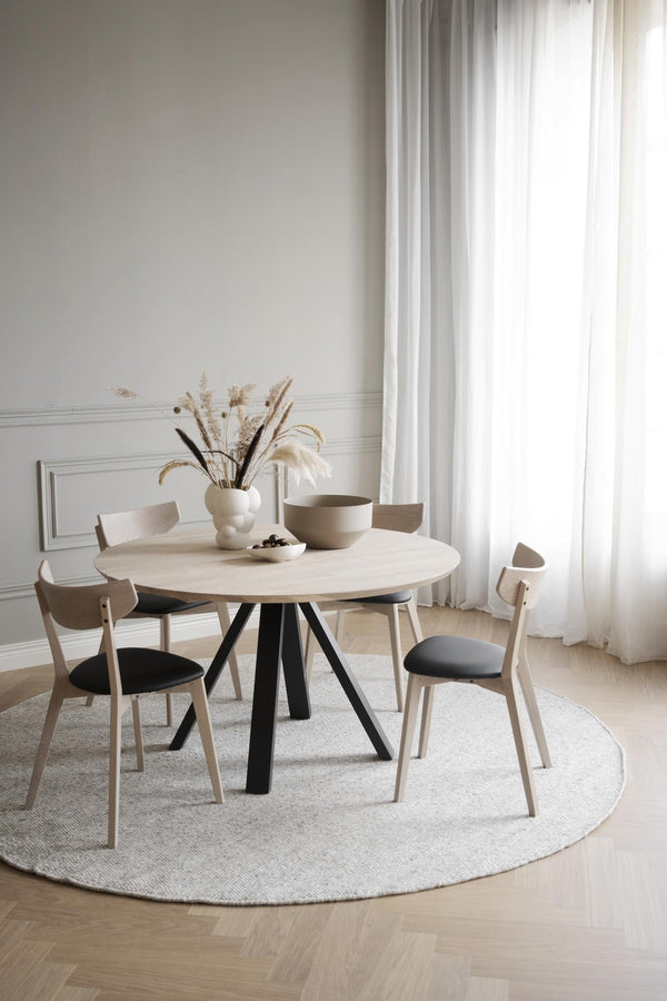 Runt matbord i ljust trä med svarta ben, omgivet av fyra stolar med grå dynor på ljus rund matta