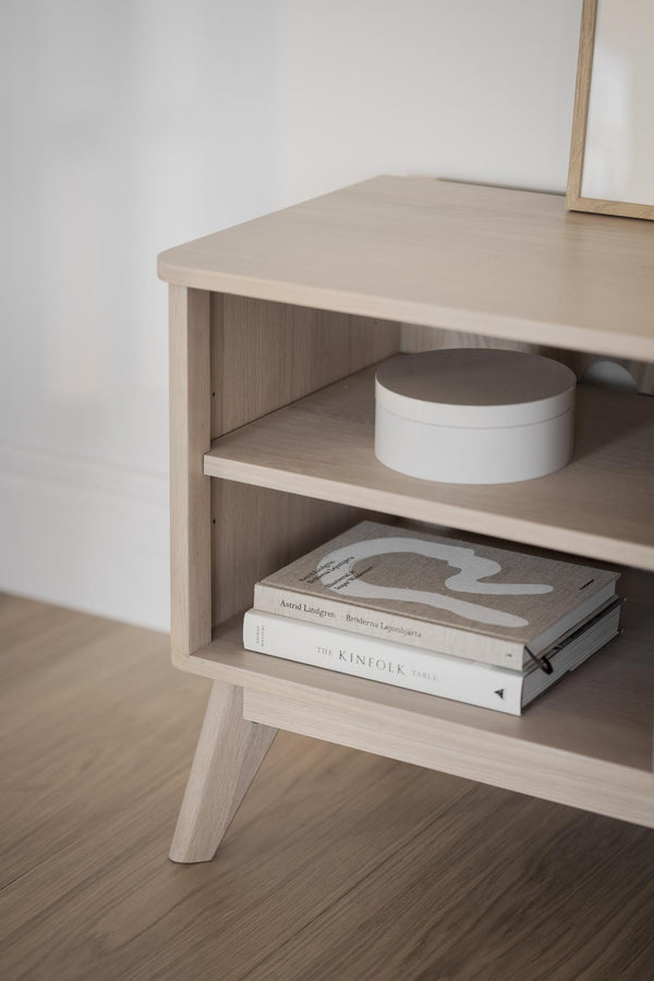 Sängbord i ljust trä med två öppna hyllplan, dekorerat med böcker och en vit ask