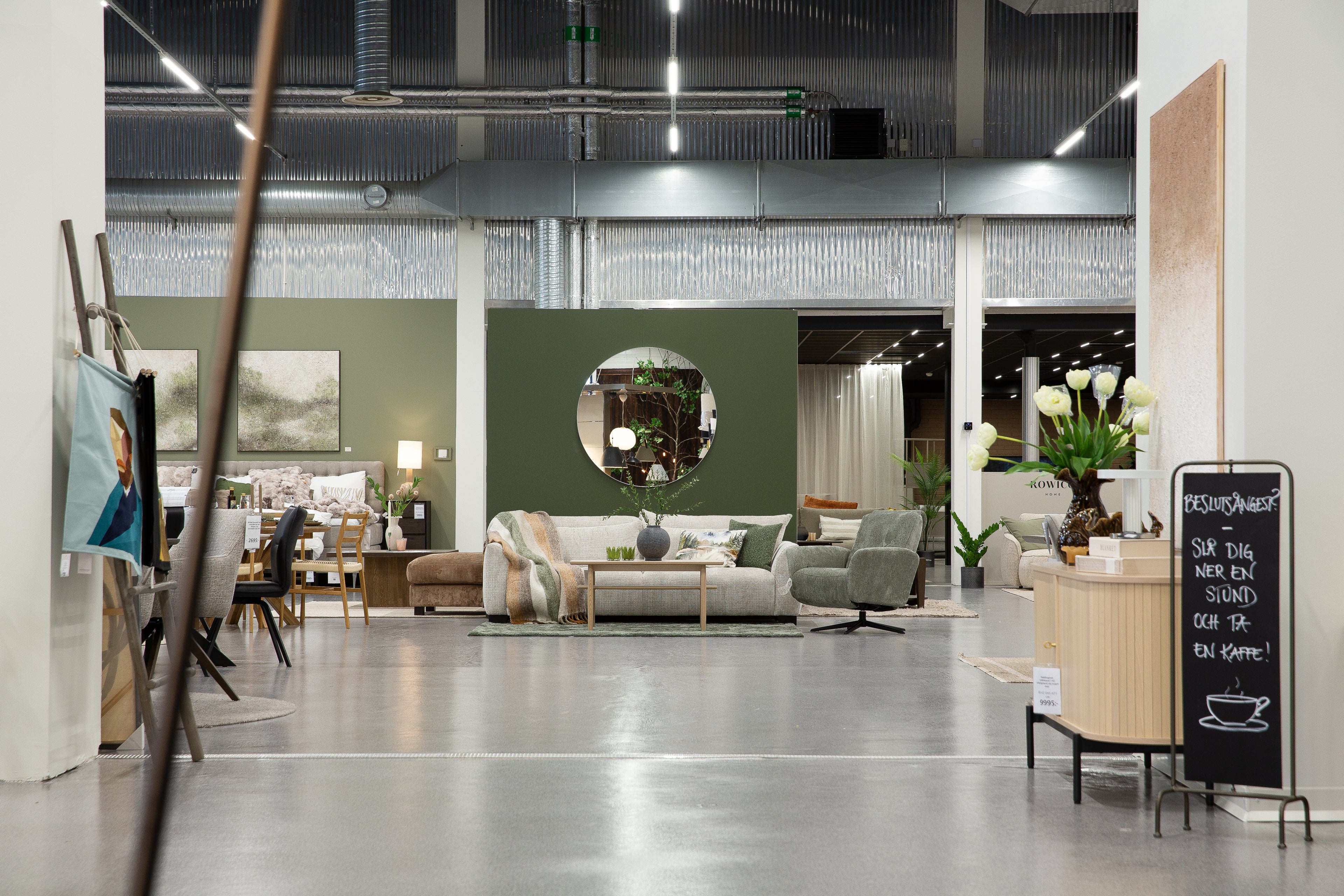 Möbelaffär med industriell inredning, gröna väggar, soffa, fåtöljer och dekorativa växter i showroom