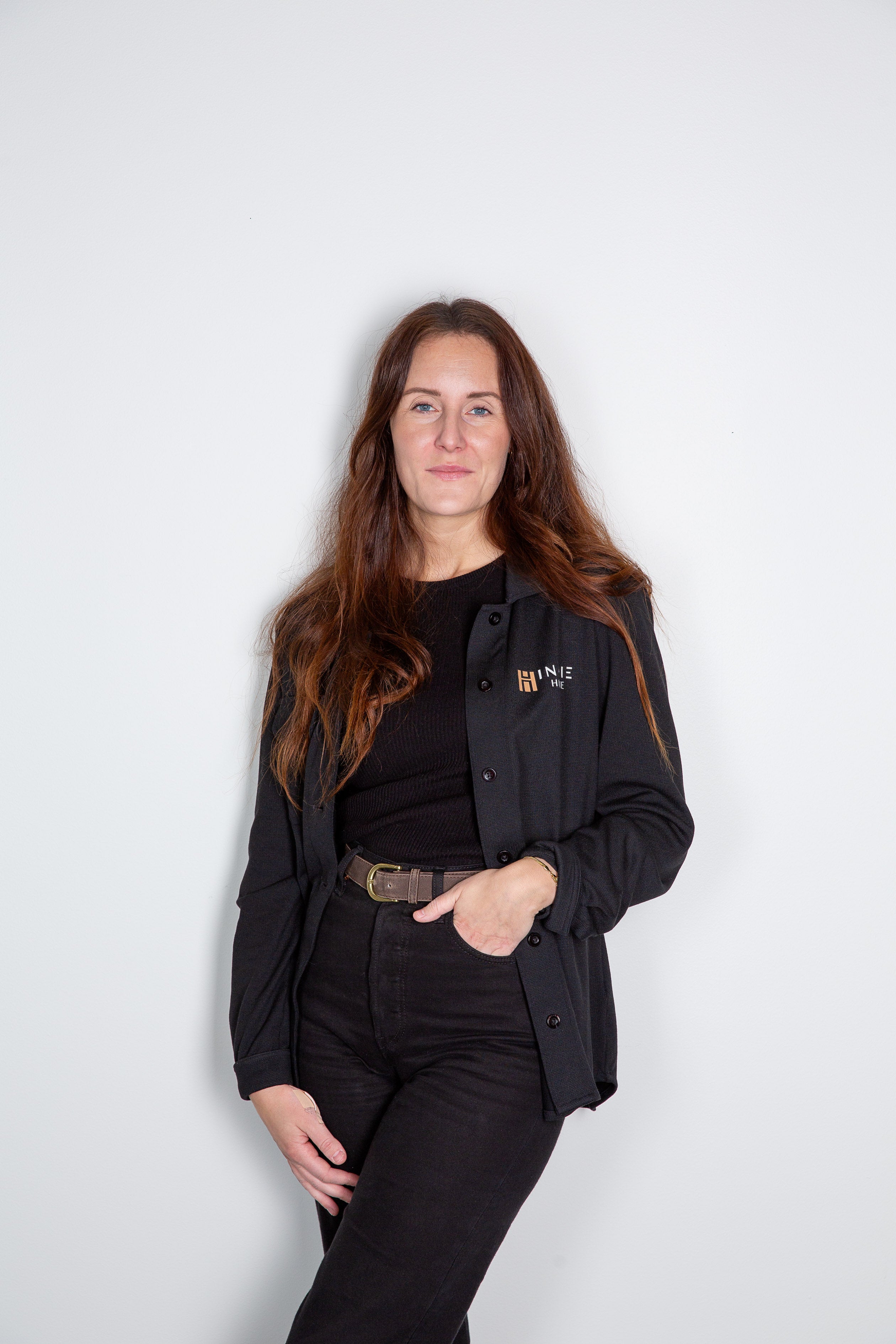 Kvinna i svart kavaj med logotyp, svart tröja och svarta jeans med brunt skärp