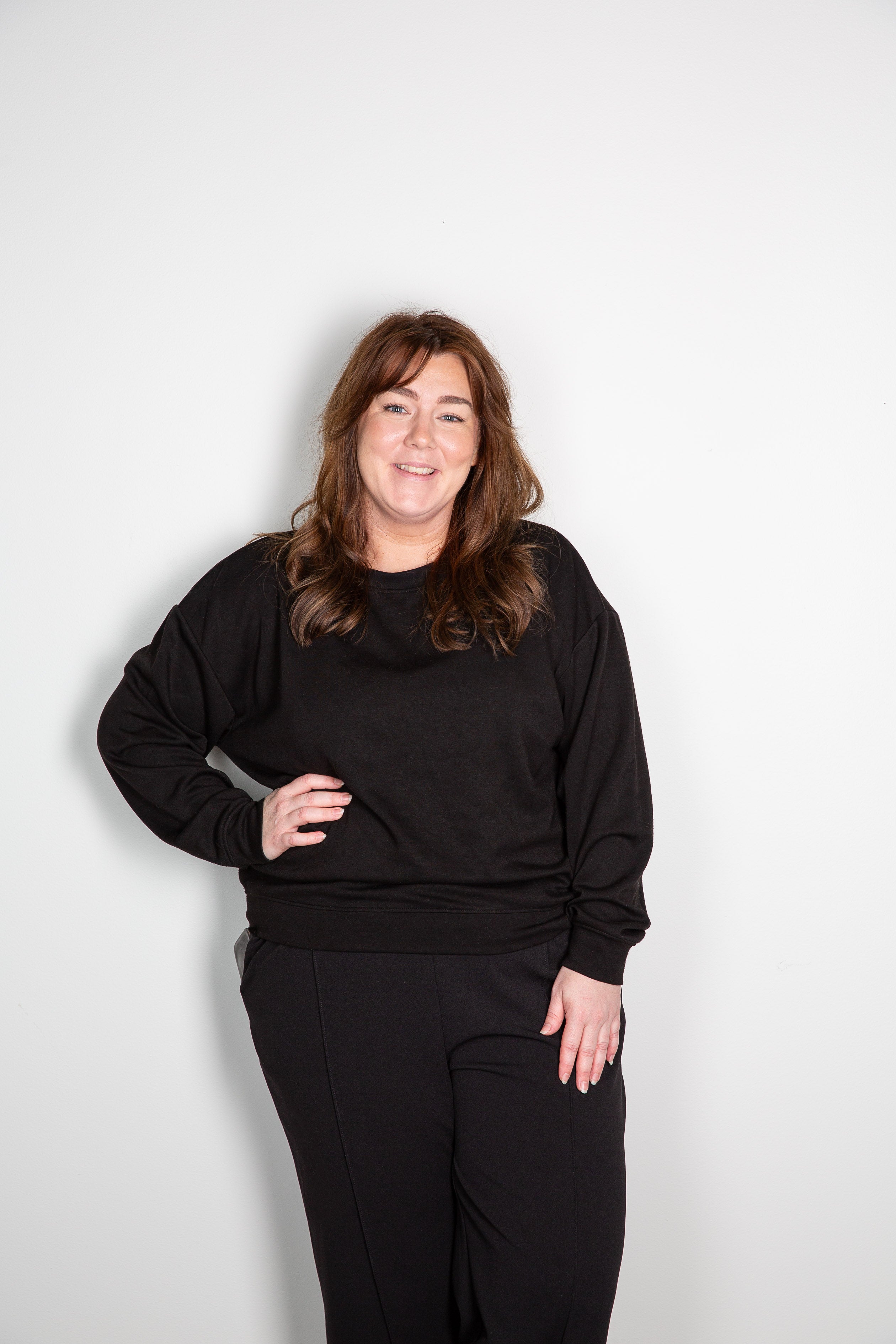 Kvinna i svart oversized sweatshirt och svarta byxor poserar mot vit bakgrund