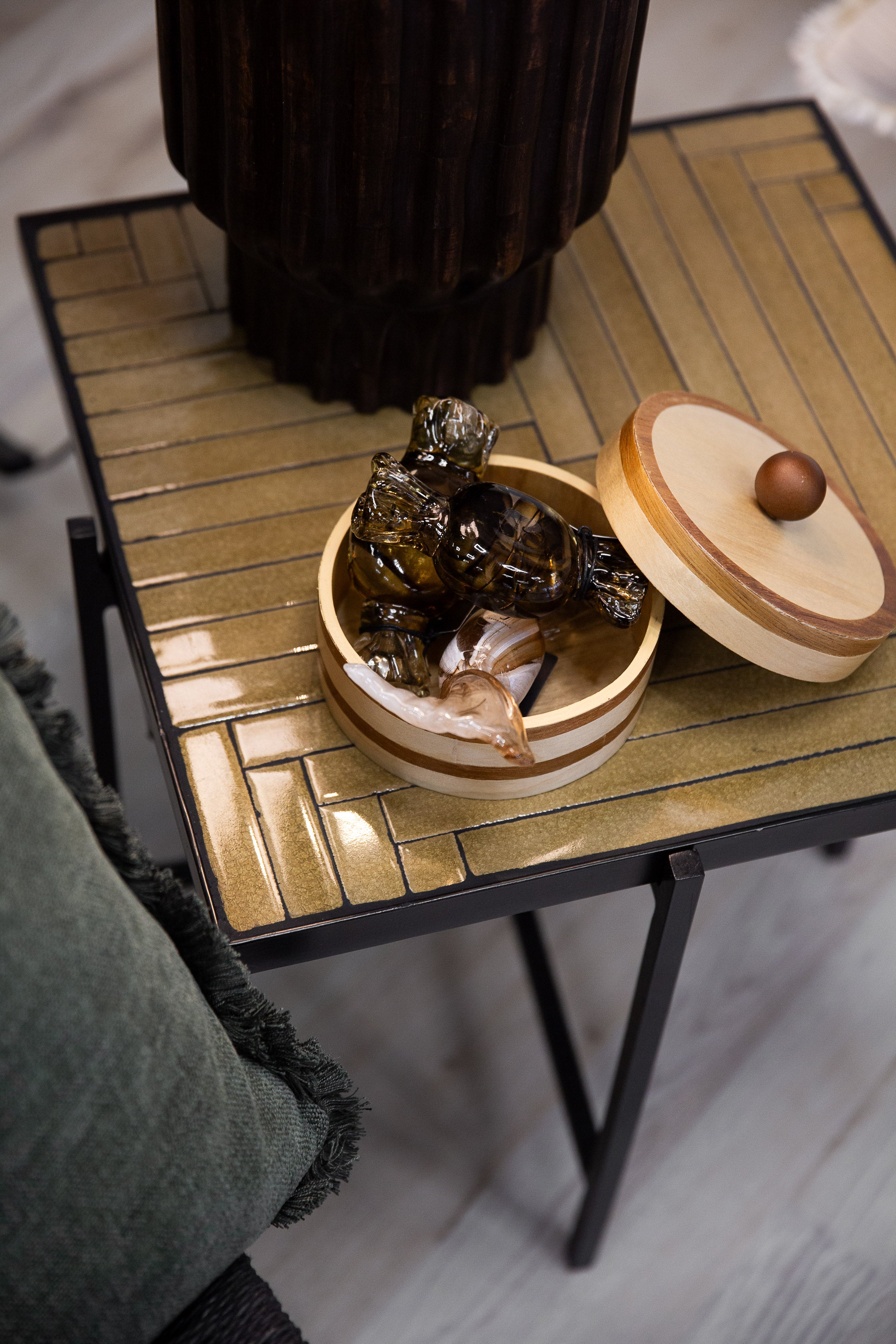 Rund förvaringsbox i trä med lock och dekorativ knopp på gyllene bricka bredvid mörk vas