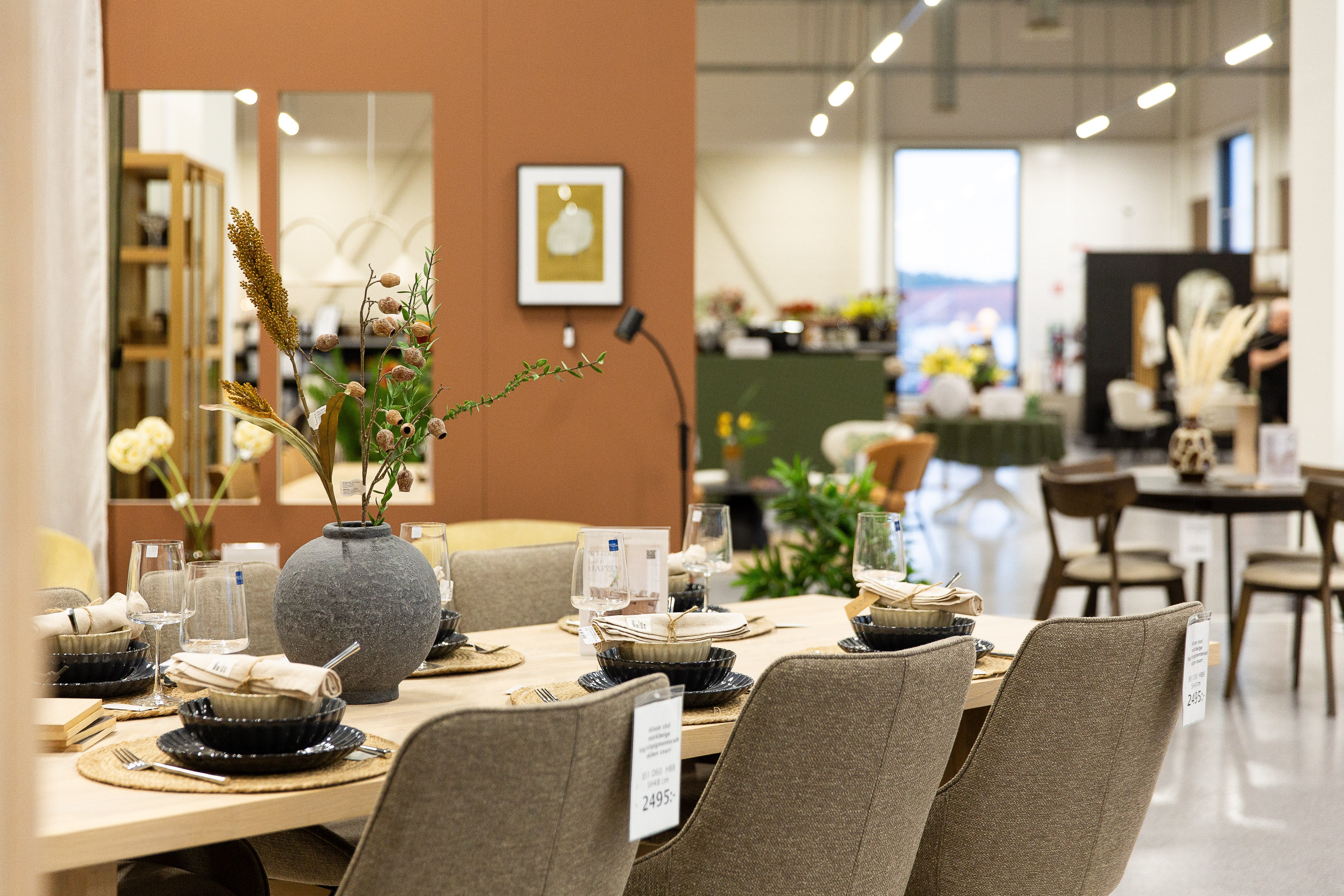 Matbord i ljust trä med gråa stolar, uppdukat med tallrikar, glas och vas med torkade blommor i showroom