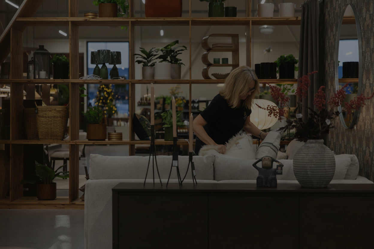 Kvinna sitter i modern soffa omgiven av växter och inredningsdetaljer i ljust showroom med träbokhylla